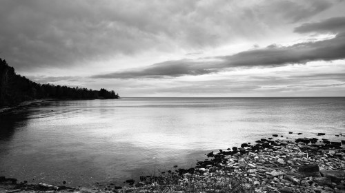 A serene black and white seascape with calm waters, rocky shore, and overcast skies.