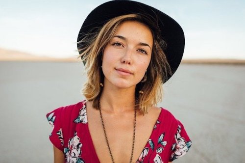 A young woman with shoulder-length hair wears a black hat and a floral dress, standing in a desert landscape.