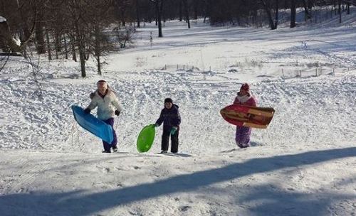 Three children climb a snowy hill, each holding a colorful sled, ready to go sledding on a winter day.
