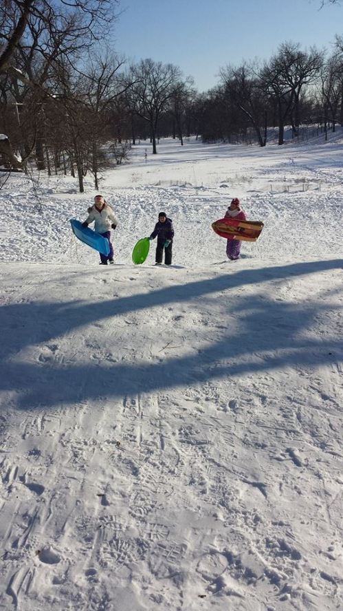 Three children climb a snowy hill, each holding a colorful sled, ready to go sledding on a winter day.