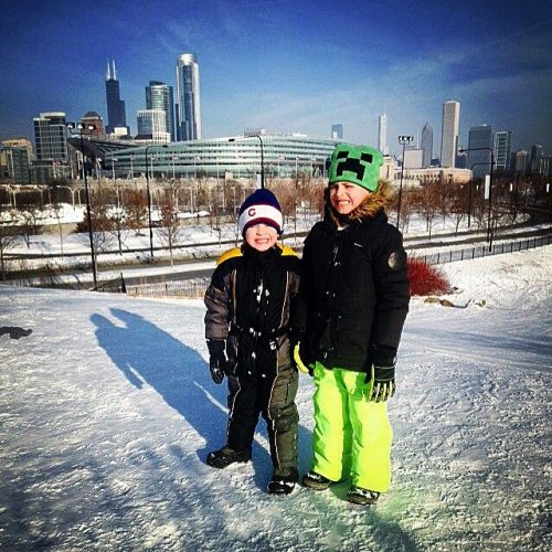 Two children stand on a snowy hill, smiling against a city skyline backdrop. One wears a winter hat and bright green pants.