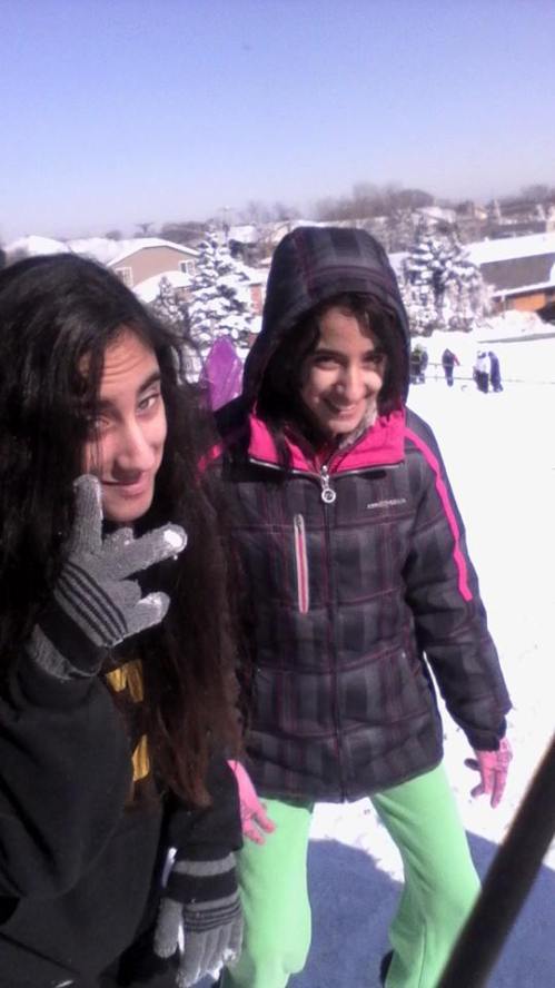 Two girls playfully pose in the snow, wearing winter jackets and gloves, with a snowy landscape in the background.