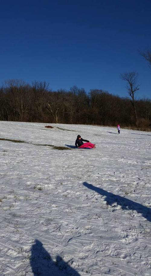 A person sleds down a snowy hill, with trees in the background and another person walking in the distance.