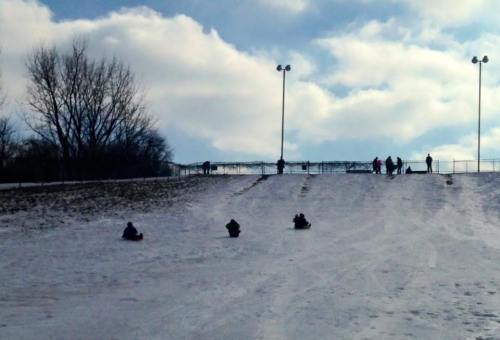 People sledding down a snowy hill under a cloudy sky, with trees and lights in the background.