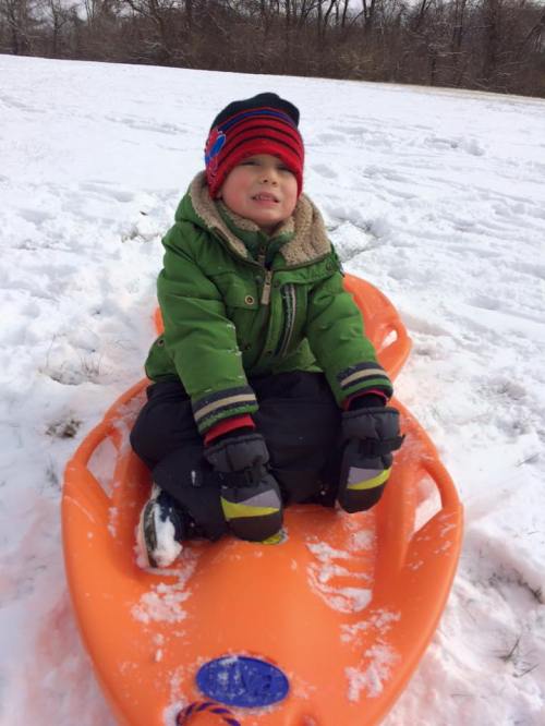 A young child in a green jacket and red hat sits on an orange sled in a snowy field, smiling happily.