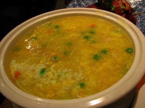 A bowl of yellow soup with green peas and bits of vegetables, served in a white ceramic dish.