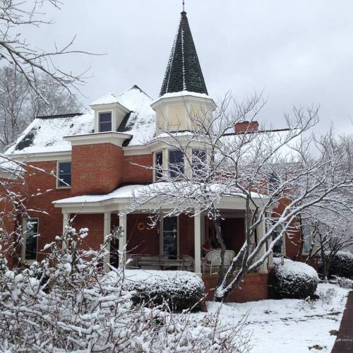 A snow-covered brick house with a pointed roof and a porch, surrounded by bare trees in winter.