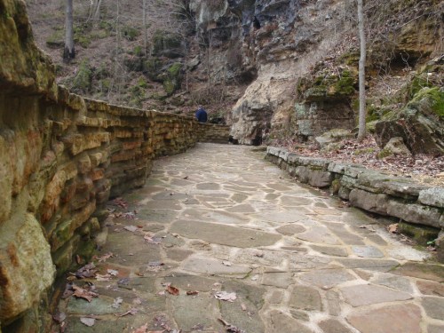 A stone pathway winds through a wooded area, flanked by rocky cliffs and scattered leaves. A person is visible in the distance.