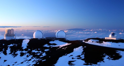 Three observatories on a snow-covered mountain peak, surrounded by clouds under a blue sky at sunset.