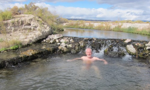 A man relaxes in a natural hot spring, surrounded by rocks and greenery, with a cloudy sky above.
