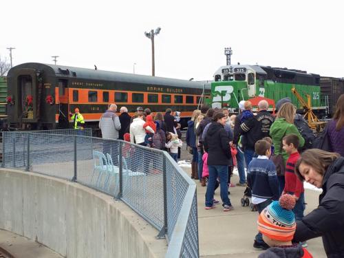 A crowd gathers near a vintage train and locomotive, decorated for the holidays, with families and children enjoying the event.