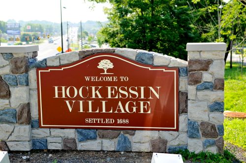 Welcome sign for Hockessin Village, established in 1688, surrounded by greenery and a road in the background.