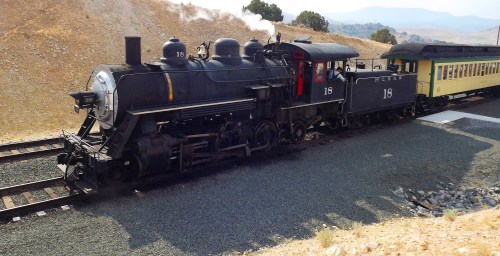 A vintage steam locomotive with a black engine and yellow passenger car, parked on a gravel track in a rural setting.
