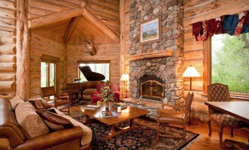 Cozy log cabin interior featuring a stone fireplace, piano, and rustic furniture with large windows and natural light.