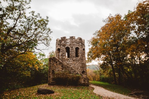 A stone tower surrounded by trees with autumn foliage, set against a cloudy sky. A path leads to the tower.