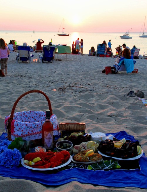A beach picnic at sunset, featuring a spread of seafood, fruits, and drinks, with people enjoying the view in the background.