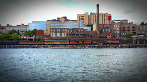 A train travels along a waterfront, with buildings and a smokestack in the background under a cloudy sky.
