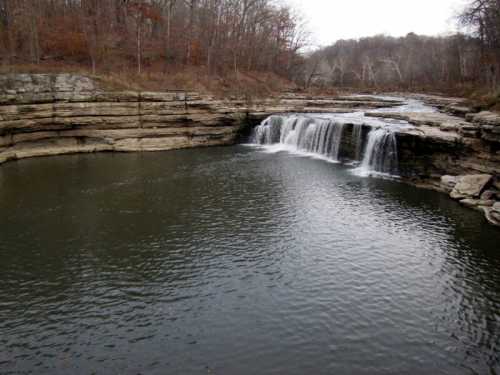 A serene waterfall cascades over rocky cliffs into a calm pool, surrounded by bare trees and a gray sky.