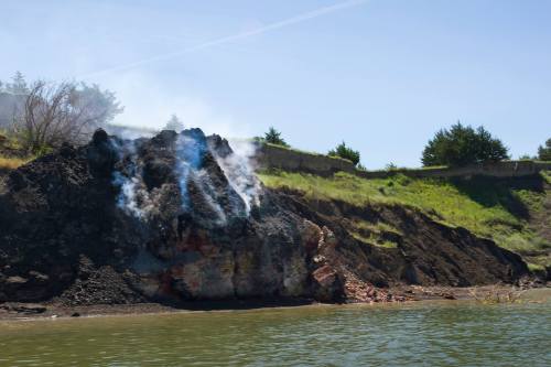 A hillside with smoke rising from a rocky outcrop near a body of water, surrounded by green vegetation.