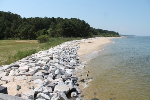 A rocky shoreline curves along a sandy beach, bordered by green trees and calm water under a clear blue sky.