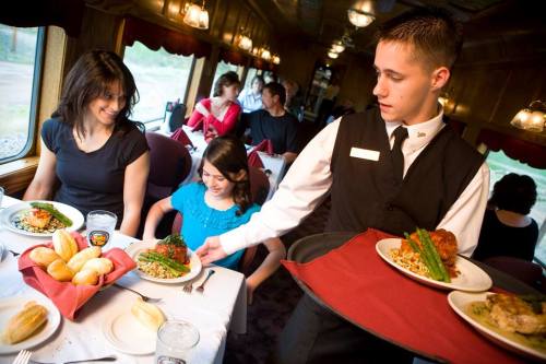 A waiter serves a meal to a woman and a girl at a dining table in a train restaurant, with other diners in the background.