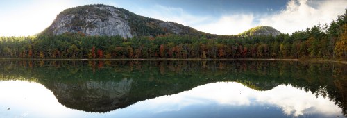 A serene lake reflects lush green hills and autumn foliage under a clear blue sky.