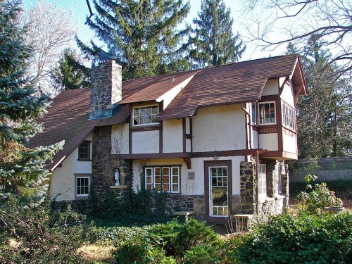 A charming two-story house with a stone chimney, surrounded by trees and greenery.