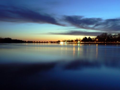 A tranquil river at dusk, reflecting colorful clouds and lights from the shore under a deep blue sky.