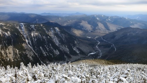 A panoramic view of snow-covered mountains and valleys under a cloudy sky, with a winding road visible in the distance.