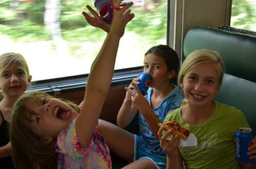 Four children enjoying snacks and drinks on a train, with one girl playfully raising her hands in excitement.