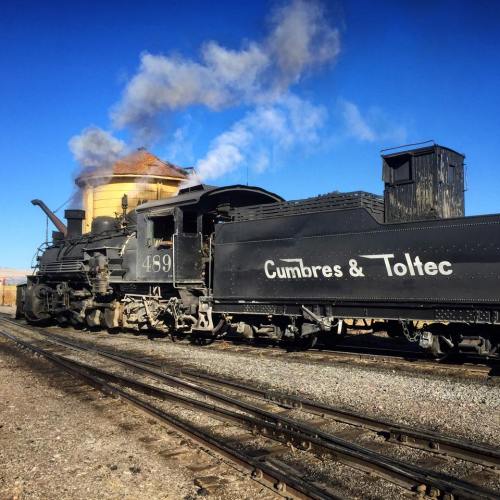 A vintage steam locomotive labeled "Cumbres & Toltec" with smoke billowing, parked on tracks in a scenic landscape.