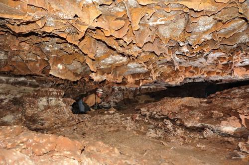 A person exploring a cave with textured, orange-brown rock formations and stalactites overhead.