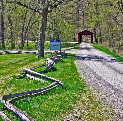 A gravel road leads to a red covered bridge, surrounded by trees and a wooden fence in a scenic landscape.