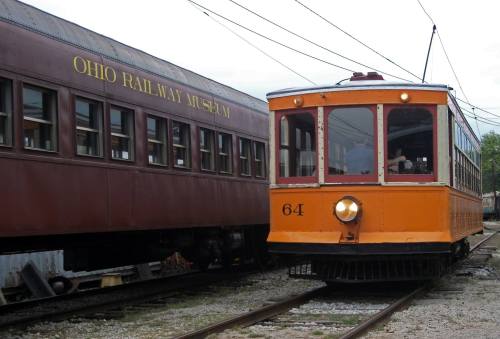 An orange vintage streetcar numbered 64 next to a brown train car labeled "Ohio Railway Museum."