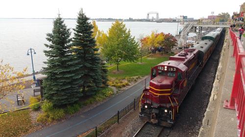 A train approaches a lakeside station, surrounded by trees and autumn foliage, with people gathered nearby.