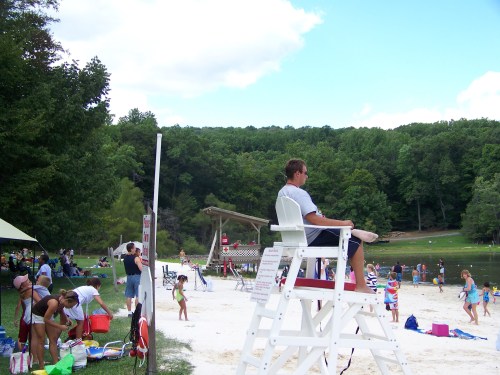 A lifeguard sits on a tall chair overlooking a busy beach with people swimming and relaxing by a lake surrounded by trees.