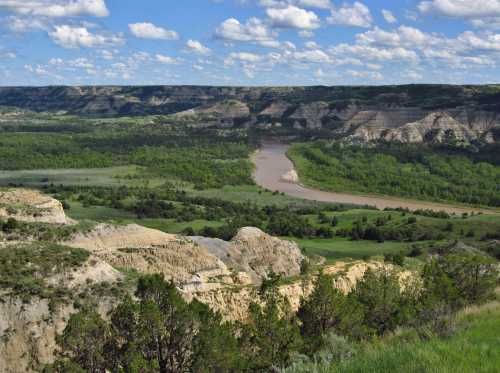 A scenic view of a river winding through green valleys and rugged hills under a blue sky with fluffy clouds.