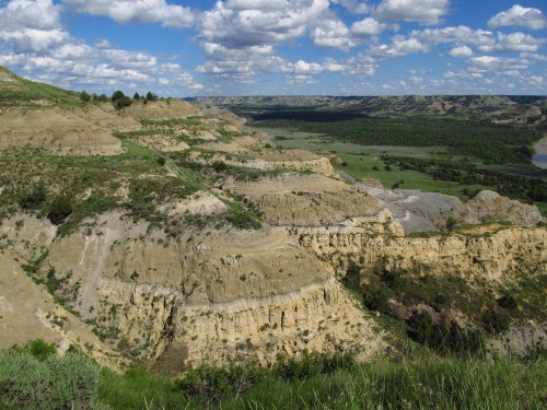 A scenic view of layered hills and valleys under a blue sky with fluffy clouds, showcasing lush greenery and rugged terrain.