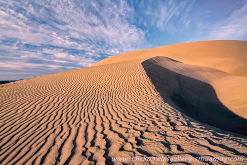 A sandy dune with rippled patterns under a blue sky with wispy clouds, creating a serene desert landscape.