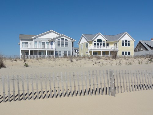 Two beach houses stand on a sandy shore, separated by a wooden fence, under a clear blue sky.