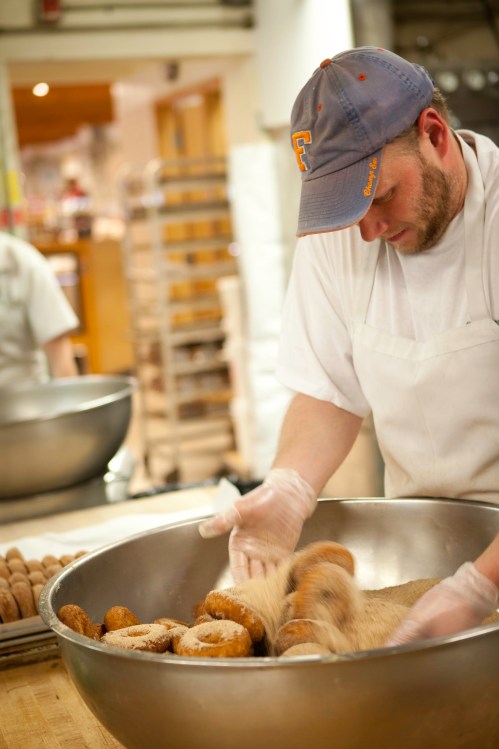 A baker in a white apron and cap mixes freshly made donuts in a large bowl, preparing them for coating.