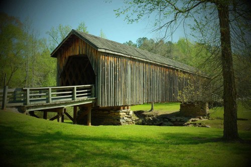 A rustic wooden covered bridge spans a small stream, surrounded by lush greenery and trees.