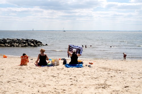 Three people relax on a sandy beach, with two sitting on towels and one playing in the water under a cloudy sky.