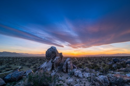 A rocky landscape under a vibrant sunset, with colorful clouds and distant mountains in the background.