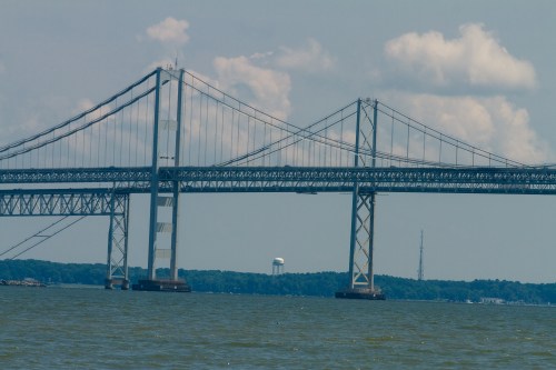 A large suspension bridge spans a body of water, with clouds in the sky and a distant water tower visible.