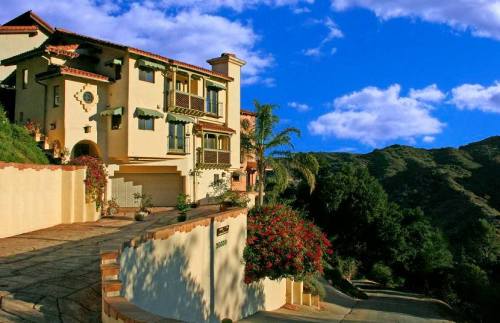 A beautiful hillside home with a Mediterranean style, surrounded by lush greenery and a blue sky with clouds.