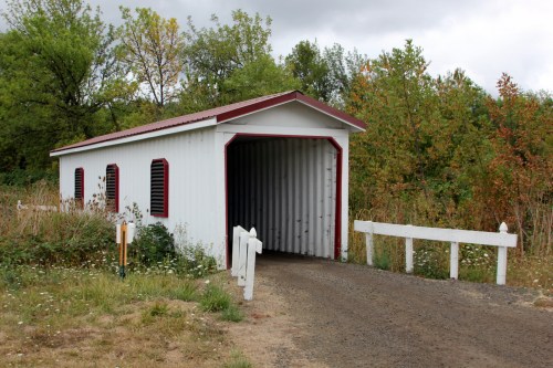 A white covered bridge with red accents, surrounded by greenery and a gravel path leading to it.