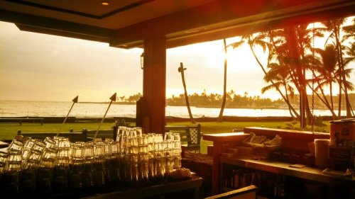 A beachside bar with stacked glasses, palm trees, and a sunset over the ocean in the background.