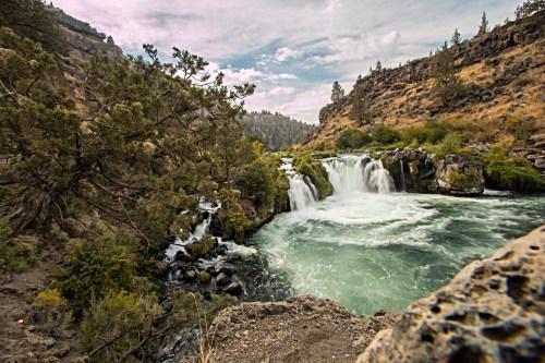 A scenic view of a waterfall cascading into a turquoise river, surrounded by rocky cliffs and lush greenery.