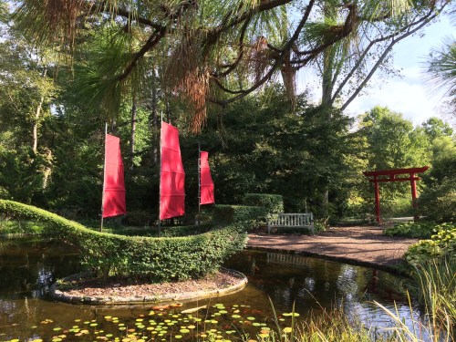 A serene garden scene featuring red sails, lush greenery, a pond, and a traditional red archway.